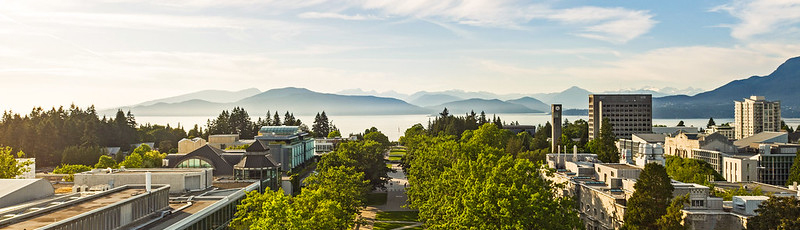 Aerial view of Main Mall facing north, with trees and buildings lining the main walkway, water and mountains at the horizon under a blue sky with wispy clouds.