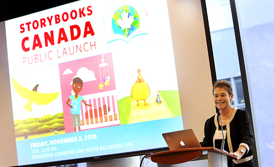 Bonny Norton is standing at a podium, with an image projected on a screen behind her, which reads: Storybooks Canada Public Launch. Friday, November 2, 2018. 3:00 - 6:00 pm. Ponderosa Commons Oak House Ballroom, UBC. With illustrations of a bird flying over a hilly landscape and people, a child holding a book leaning toward a baby in a crib with a mobile above, and a chicken walking beside an antennaed, unidentified animal.