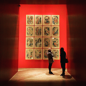 Two women standing in front of a spotlighted gallery installment with 12 images on the wall.