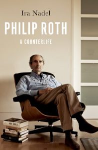 Book cover for Philip Roth: A Counterlife by Ira Nadel, with portrait of Roth reclining in a chair in a room with bare wall and a rug, beside a stack of books on the wood floor