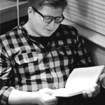 Dallas Hunt seated in a corner of a room, looking down at a book he's holding in two hands, wearing a gingham button-down top