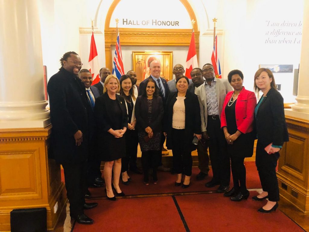 Swahili Vision International Association photo of group members standing with elected officials from the Province of British Columbia in hall with flags and "Hall of Honour" sign above them, including Premier John Horgan and Minister Anne Kang