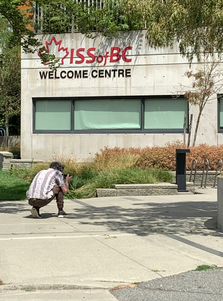 Flick Harrison is crouched, back turned, on the sidewalk outside the Immigration Services Society of BC taking b-roll footage for the Borderstory film on refugees and borders.