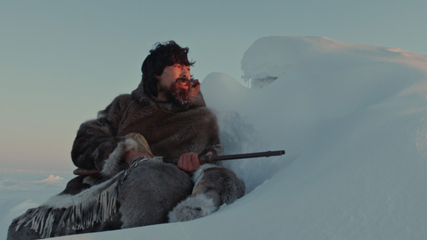 Benjamin Kunuk as Kuanana, looking over a snowdrift into the distance with sunlight on his face, seated, carrying a long gun. Still from Maliglutit via The Cinematheque website, PHOTO COURTESY KINGULIIT PRODUCTIONS INC.