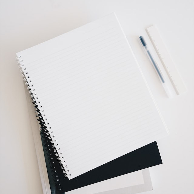 A spiral-bound notebook is opened to a blank, lined page, beside a pen and ruler.