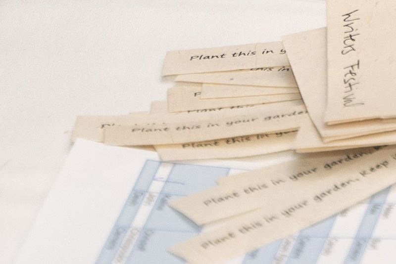 Closeup on beige slips of paper on a tabletop with words printed on them: "Plant this in your garden...", one sheet with "writers festival" written in black ink