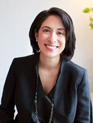 Minelle Mahtani wearing a black blazer and silver chain necklace, smiling, with a green leafy plant in the background