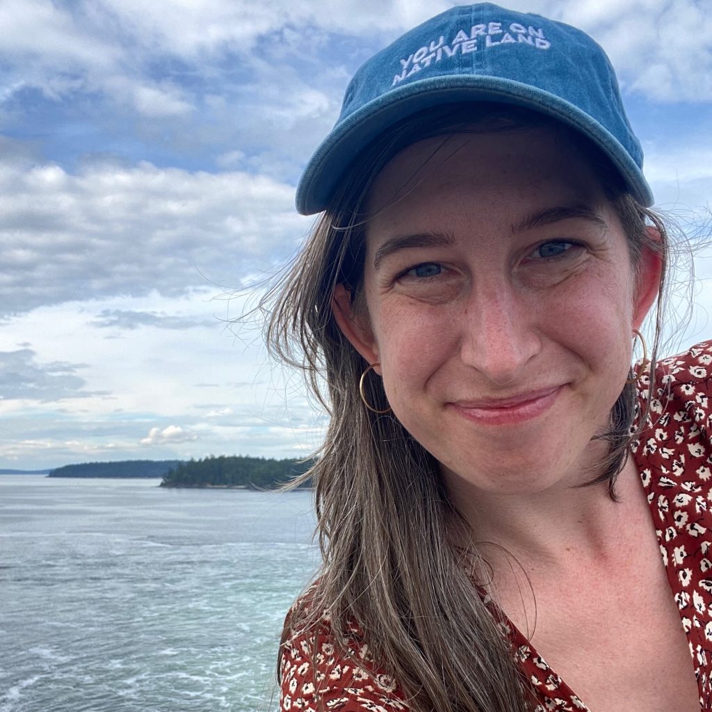 Emily Jean Leischner smiles in a photo taken outside on a boat with the ocean in the background. She has brown hair and wears a blue hat that says "You are on native land" in white letters. 