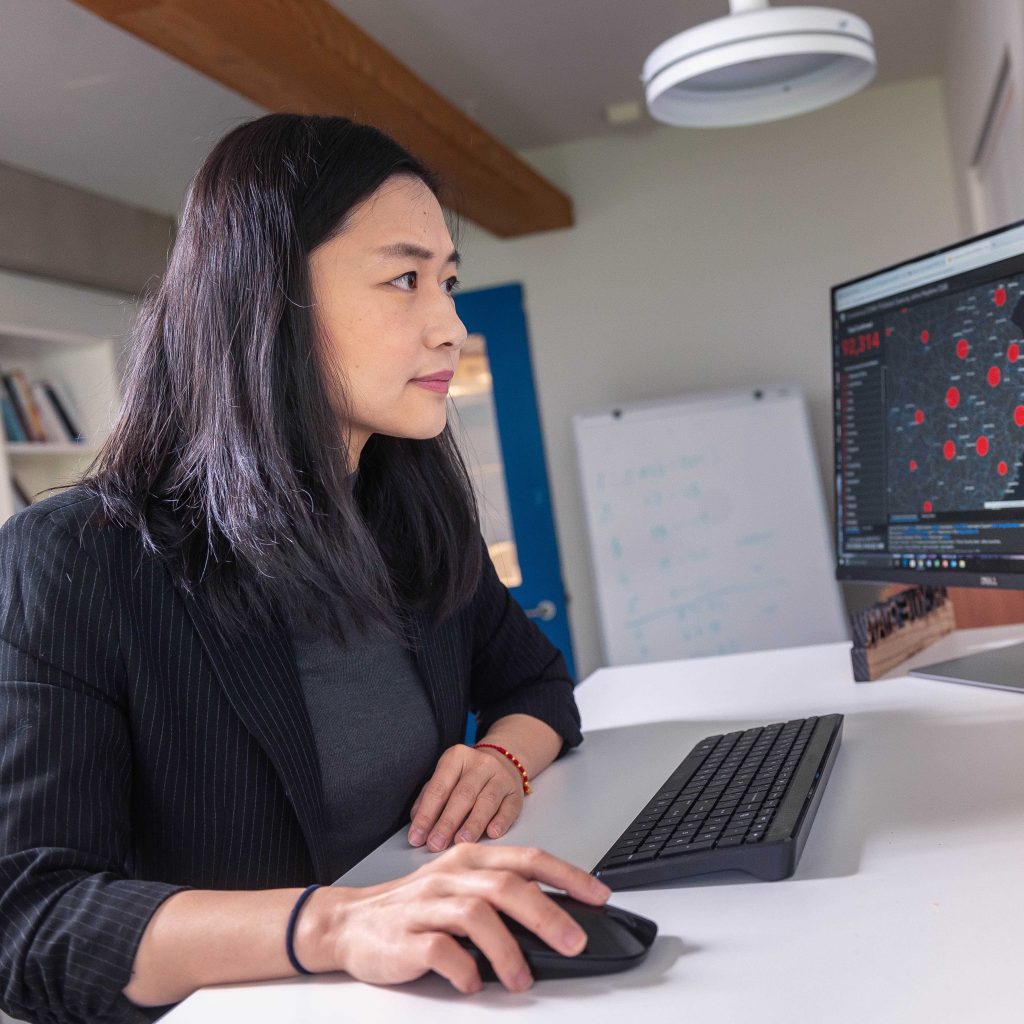 Dr. Yue Qian sits at a desk with her hand on her computer mouse looking at data on a monitor. She has black hair and is wearing a black blazer.