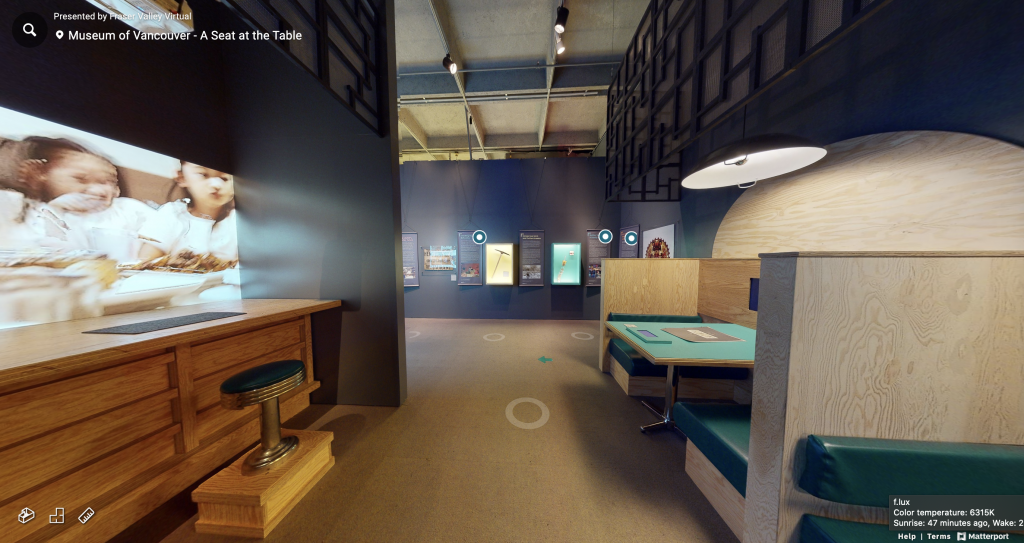 A capture of part of the virtual "A Seat at the Table" MOV exhibit. A counter can be seen on the left side of the hallway facing a screen projecting an image of children. On the right side of the hallway are table booths. Down the hallway, there is a framed pickaxe as artefact. 