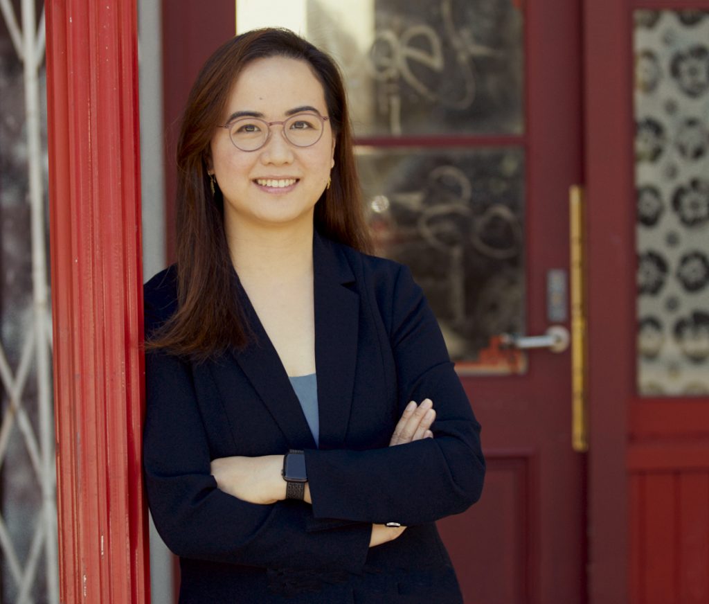Denise Fong smiles while leaning against a red door frame wearing a black blazer. She wears glasses and has brown hair.