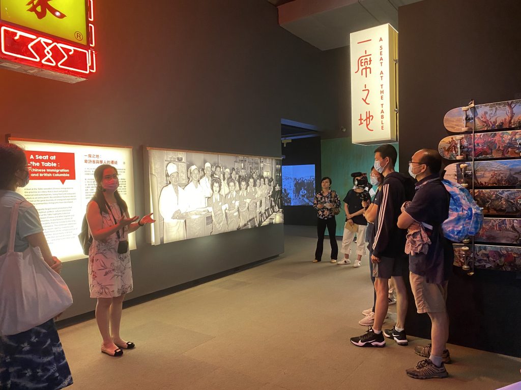 Denise Fong is talking to a group of visitors in front of photos featured as a part of the "A Seat at the Table" exhibit at MOV.
