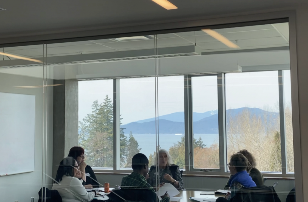 A peak into a seminar room, where five students are seated at a roundtable, listening attentively to a speaker sitting at the middle of the table talking and gesturing. The room looks out to the water and mountains. 