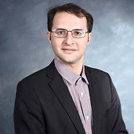 Hessam smiles softly into the camera, against a gradient gray background, wearing glasses, a black jacket, and a lavender shirt.