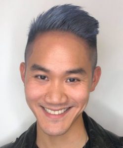 A close-up shot of David Ng smiling into the camera, hair dyed a dusty indigo, wearing a black collared shirt.