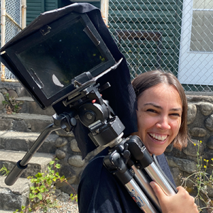 Erin with a short bob smiling into the camera, carrying videocamera equipment on her shoulder.