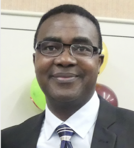 A medium shot of Joash smiling into the camera, wearing glasses and a suit.